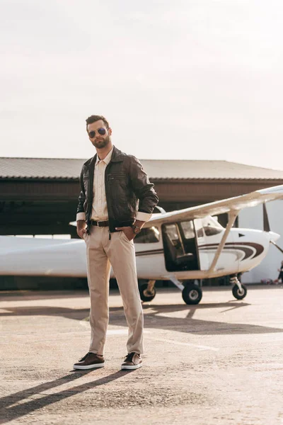 Seriöser männlicher Pilot in Lederjacke und Sonnenbrille posiert in Flugzeugnähe — Stockfoto