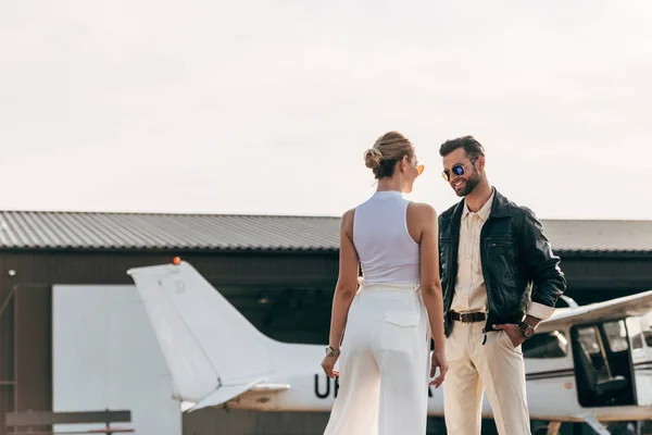 Задний вид стильной женщины разговаривающей с улыбающимся парнем в кожаной куртке и солнцезащитных очках рядом с самолетом — стоковое фото