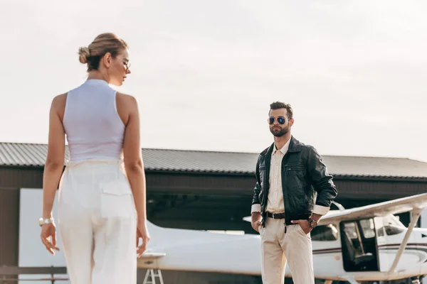 Jovem confiante em jaqueta de couro e óculos de sol olhando para namorada perto de avião — Fotografia de Stock