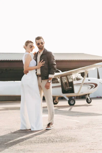 Joven guapo en chaqueta de cuero y gafas de sol abrazando novia atractiva cerca de avión - foto de stock