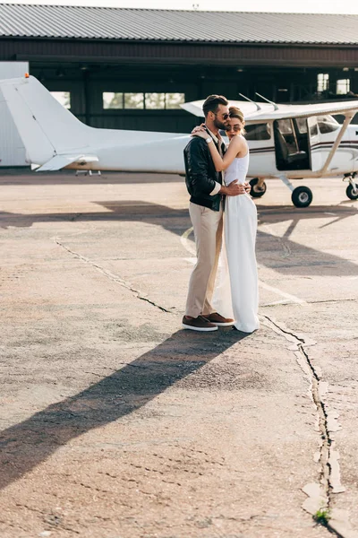 Bel homme en veste en cuir et lunettes de soleil embrassant petite amie attrayante près de l'avion — Photo de stock