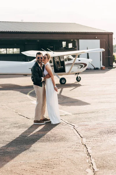 Laughing young man in leather jacket and sunglasses embracing attractive girlfriend near airplane — Stock Photo