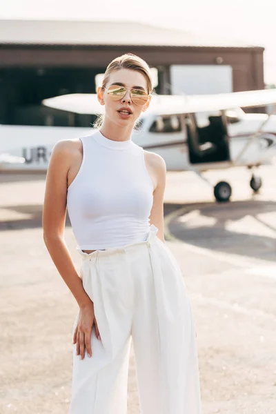 Bella giovane donna in occhiali da sole e top bianco in posa vicino aereo — Foto stock