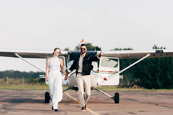 Homme souriant en veste en cuir pointant du doigt à la petite amie marchant près de l'avion — Photo de stock