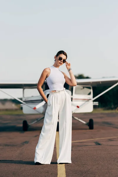 Стильна молода жінка в сонцезахисних окулярах позує біля літака — стокове фото