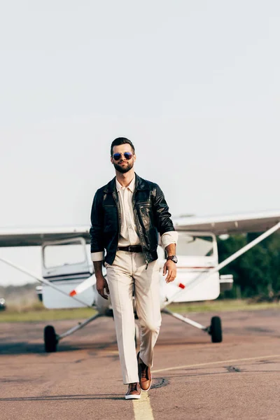 Piloto masculino guapo en chaqueta de cuero y gafas de sol caminando cerca de aviones - foto de stock