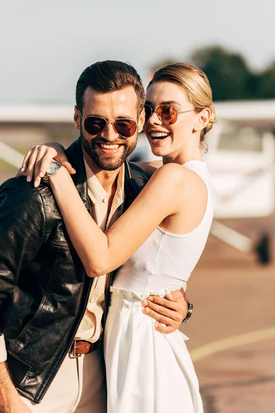 Laughing attractive woman hugging stylish boyfriend in leather jacket and sunglasses near plane — Stock Photo