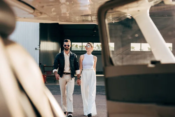 Селективное внимание стильного мужчины в кожаной куртке и солнцезащитных очках, идущего с подругой рядом с самолетом — стоковое фото