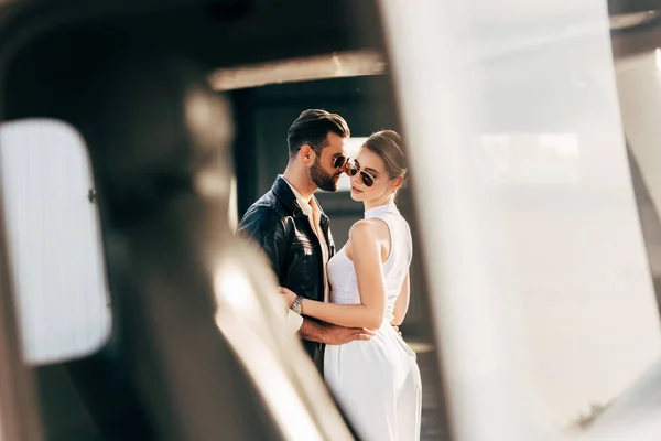 Enfoque selectivo del hombre en chaqueta de cuero y gafas de sol abrazando novia atractiva cerca de avión - foto de stock