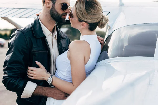 Jovem bonito em jaqueta de couro e óculos de sol abraçando namorada perto do avião — Fotografia de Stock