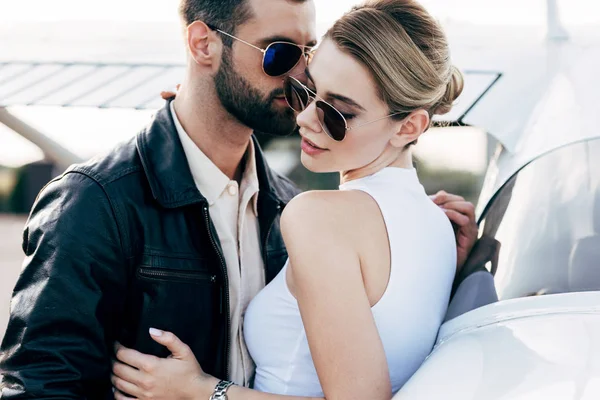 Hombre con estilo en chaqueta de cuero y gafas de sol abrazando novia atractiva cerca de avión - foto de stock