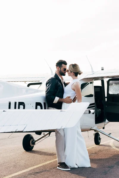 Seitenansicht von stylischem Paar mit Sonnenbrille, das sich in Flugzeugnähe umarmt — Stockfoto