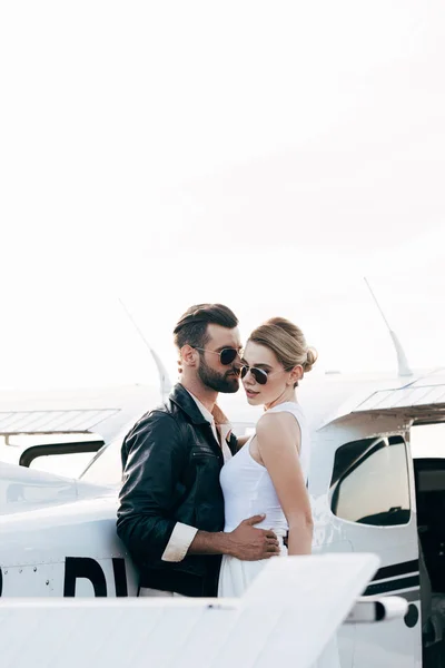 Side view of stylish man in leather jacket and sunglasses embracing attractive girlfriend near plane — Stock Photo