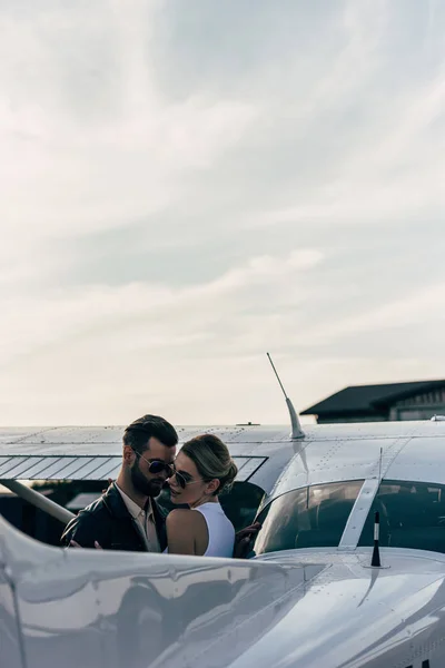 Hombre guapo serio en chaqueta de cuero y gafas de sol abrazando novia atractiva cerca de avión - foto de stock