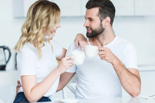 Feliz joven pareja sosteniendo tazas de café y sonriendo entre sí - foto de stock