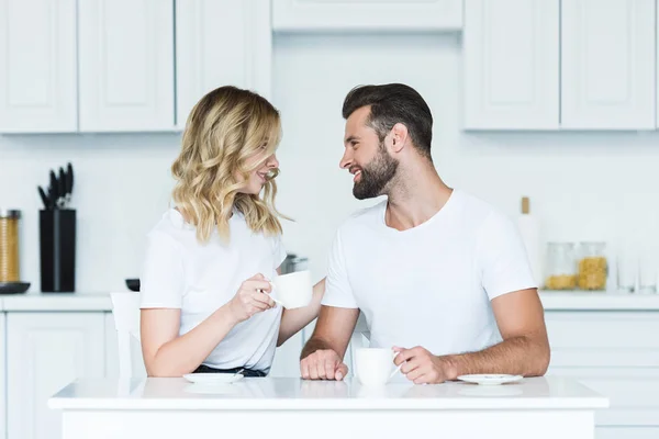 Feliz jovem casal bebendo café e sorrindo uns aos outros na cozinha de manhã — Fotografia de Stock