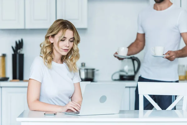 Attraente sorridente ragazza utilizzando il computer portatile mentre il fidanzato in possesso di tazze di caffè dietro — Foto stock