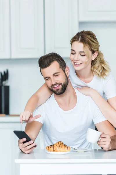 Hermosa pareja joven feliz utilizando el teléfono inteligente mientras desayunan juntos - foto de stock