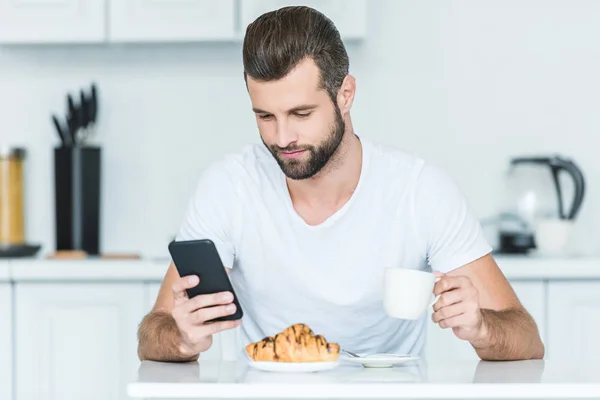 Красивий бородатий молодий чоловік використовує смартфон під час сніданку — стокове фото