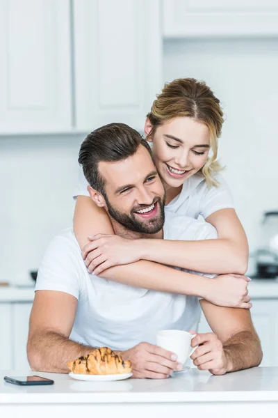 Happy young woman hugging smiling boyfriend holding cup of coffee at morning — Stock Photo