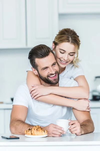 Feliz joven mujer abrazando sonriente novio sosteniendo taza de café por la mañana - foto de stock