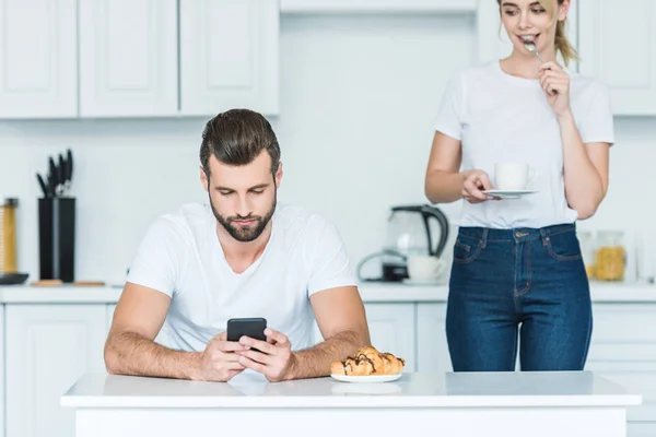 Jovem homem usando smartphone enquanto namorada segurando xícara de café atrás — Fotografia de Stock