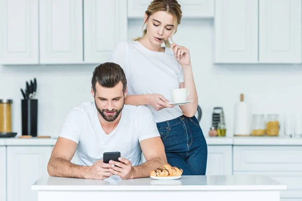 Atractiva mujer joven sosteniendo la taza de café y mirando al novio usando el teléfono inteligente - foto de stock