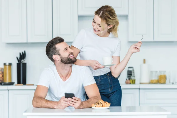 Счастливый молодой человек пользуется смартфоном и смотрит на улыбающуюся подругу, пьющую кофе по утрам — стоковое фото
