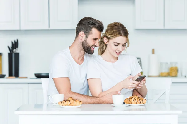 Hermosa pareja joven feliz utilizando el teléfono inteligente mientras desayunan juntos — Stock Photo