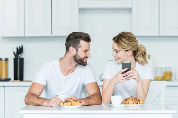 Feliz jovem casal sorrindo uns aos outros e usando smartphone enquanto tomando café da manhã juntos — Fotografia de Stock