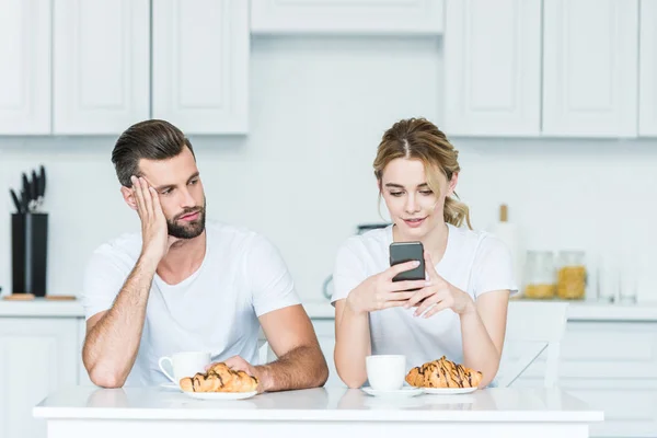 Ennuyé homme regardant sourire petite amie en utilisant smartphone pendant le petit déjeuner — Photo de stock