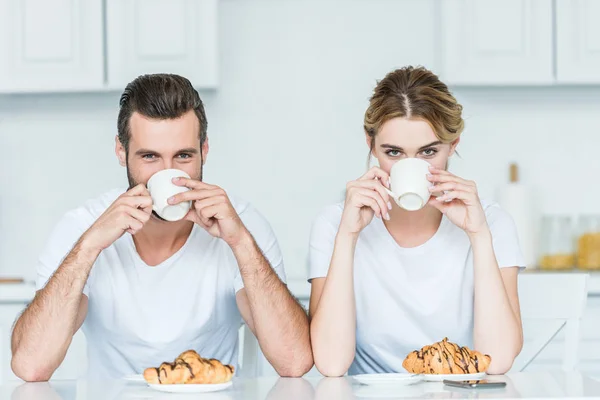 Joven pareja bebiendo café y mirando a la cámara mientras desayuna con café y croissants - foto de stock