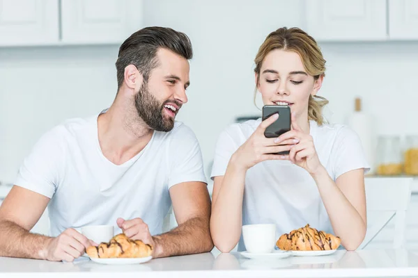 Joven sonriente mirando a la novia usando el teléfono inteligente durante el desayuno - foto de stock