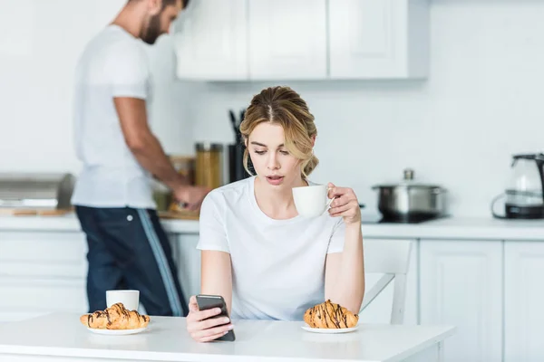 Joven mujer beber café y el uso de teléfono inteligente mientras que el novio cocinar el desayuno detrás - foto de stock