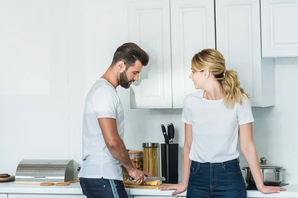 Bela jovem mulher olhando para namorado cortando baguete na cozinha — Fotografia de Stock