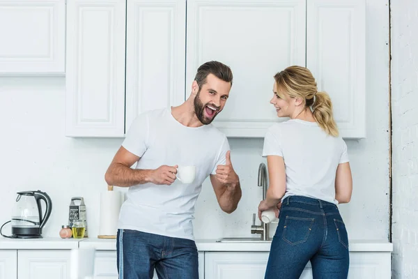 Alegre joven pareja riendo juntos en cocina, hombre mostrando el pulgar hacia arriba y sonriendo a la cámara - foto de stock