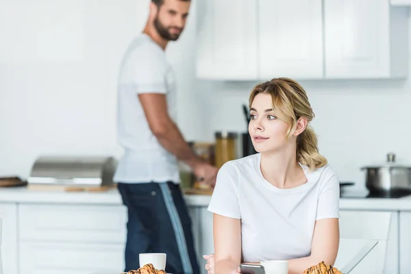 Hermosa mujer joven utilizando el teléfono inteligente y mirando hacia otro lado mientras el novio preparando el desayuno detrás - foto de stock