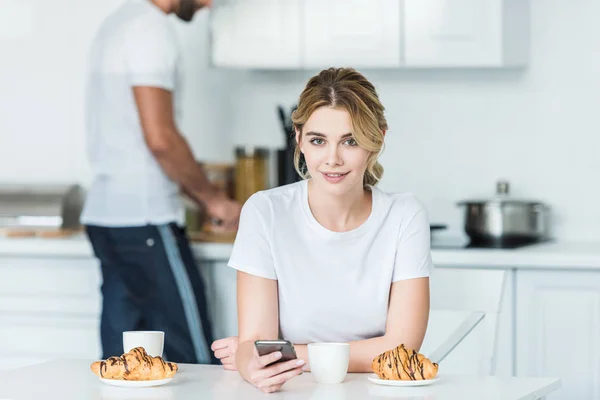 Красивая молодая женщина с помощью смартфона и улыбаясь в камеру в то время как парень готовит завтрак позади — стоковое фото