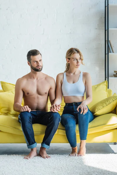 Hermosa pareja joven tomados de la mano y mirando hacia otro lado mientras se sientan juntos en el sofá amarillo - foto de stock