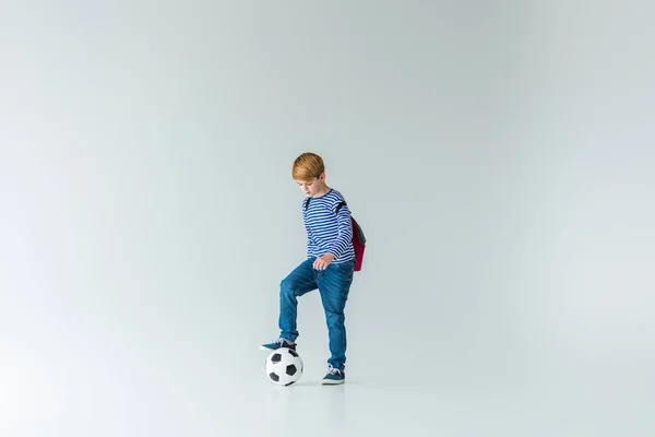 Adorável estudante com mochila colocando perna na bola de fotball no branco — Fotografia de Stock