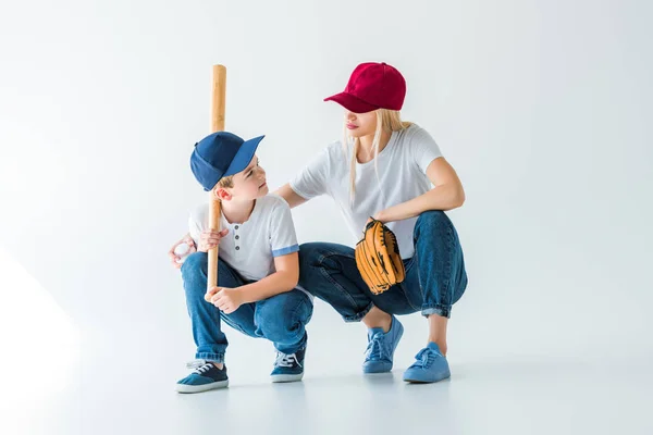 Мама и сын сидят на корточках с бейсбольной битой и перчаткой на белом и смотрят друг на друга. — стоковое фото