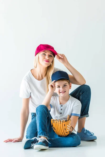 Mãe e filho tocando bonés e sentado no chão com luva de beisebol no branco — Fotografia de Stock