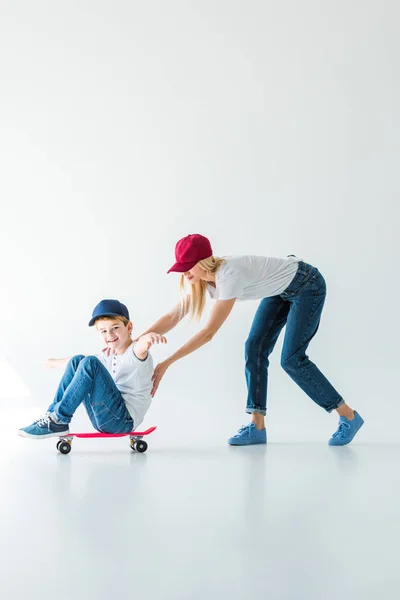 Madre in berretto rosso spingendo sorridente figlio su skate su bianco — Foto stock