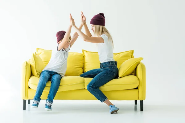 Mother and son in burgundy hats giving high fives on yellow sofa on white — Stock Photo