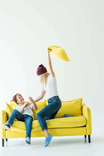 Mãe se divertindo e batendo filho com travesseiro no sofá amarelo no branco — Fotografia de Stock