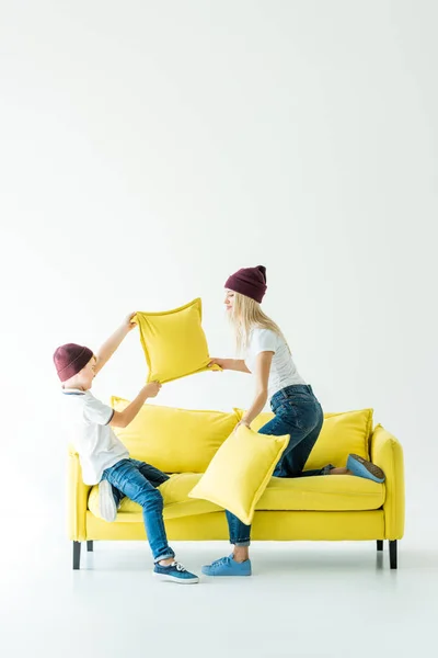 Mère et fils en chapeaux bordeaux s'amuser et se battre avec des oreillers sur canapé jaune sur blanc — Photo de stock