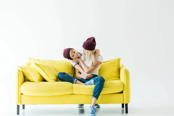 Mutter und Sohn in weinroten Hüten umarmen sich auf gelbem Sofa auf weißem Grund — Stockfoto