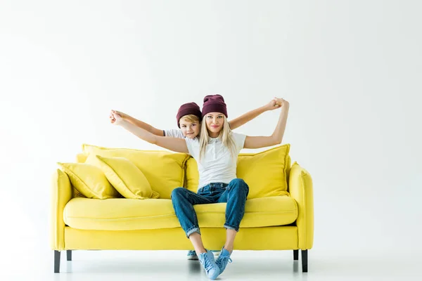 Мать и сын в бордовых шляпах притворяются, что летают на жёлтом диване на белом — стоковое фото
