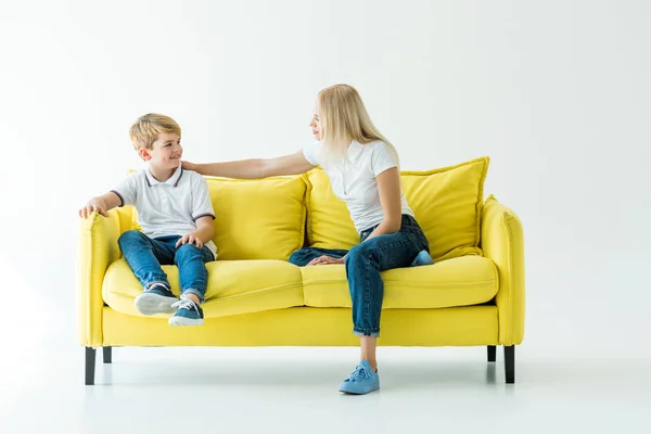 Madre e hijo sentados en un sofá amarillo y mirándose el uno al otro en blanco - foto de stock