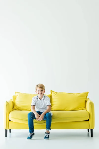 Sonriente adorable chico sentado en amarillo sofá y mirando a la cámara en blanco - foto de stock